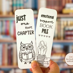 Peut inclure: Deux marque-pages rectangulaires blancs avec des illustrations et du texte en noir. Le marque-page de gauche montre un hibou lisant un livre avec le texte "just one more CHAPTER". Celui de droite représente une personne appuyée sur une pile de livres avec "emotional support BOOK PILE" et cinq petits cœurs.