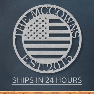 May include: A silver metal wall art piece featuring a circular design with the American flag in the center. The text "THE MCCOWNS EST. 2015" is cut out around the flag. The piece is displayed against a dark blue background.