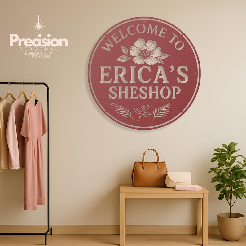 May include: A circular, dark red metal sign featuring the text "WELCOME TO ERICA'S SHESHOP" in white lettering. The design includes a central flower illustration and decorative leaf accents at the bottom, with a thin white border around the edge.