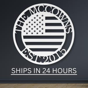 May include: White metal sign featuring a circular design with the text "THE MCCOWNS" at the top and "EST. 2015" at the bottom. The center displays an American flag design. The sign is mounted on a dark gray wall.