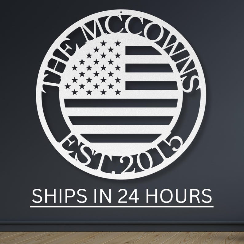 May include: White metal sign featuring a circular design with the text "THE MCCOWNS" at the top and "EST. 2015" at the bottom. The center displays an American flag design. The sign is mounted on a dark gray wall.