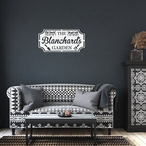 K&ouml;nnte beinhalten: Ein schwarz-wei&szlig; gemustertes Sofa mit zwei Kissen und einem Holz-Couchtisch davor. Ein wei&szlig;es Schild mit schwarzem Text lautet "The Blanchard's Garden" mit einer Schaufel- und Hacke-Grafik. Das Schild ist an einer dunkelgrauen Wand montiert.