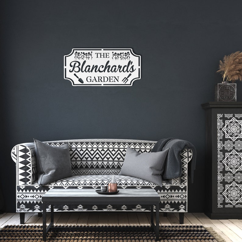 K&ouml;nnte beinhalten: Ein schwarz-wei&szlig; gemustertes Sofa mit zwei Kissen und einem Holz-Couchtisch davor. Ein wei&szlig;es Schild mit schwarzem Text lautet "The Blanchard's Garden" mit einer Schaufel- und Hacke-Grafik. Das Schild ist an einer dunkelgrauen Wand montiert.
