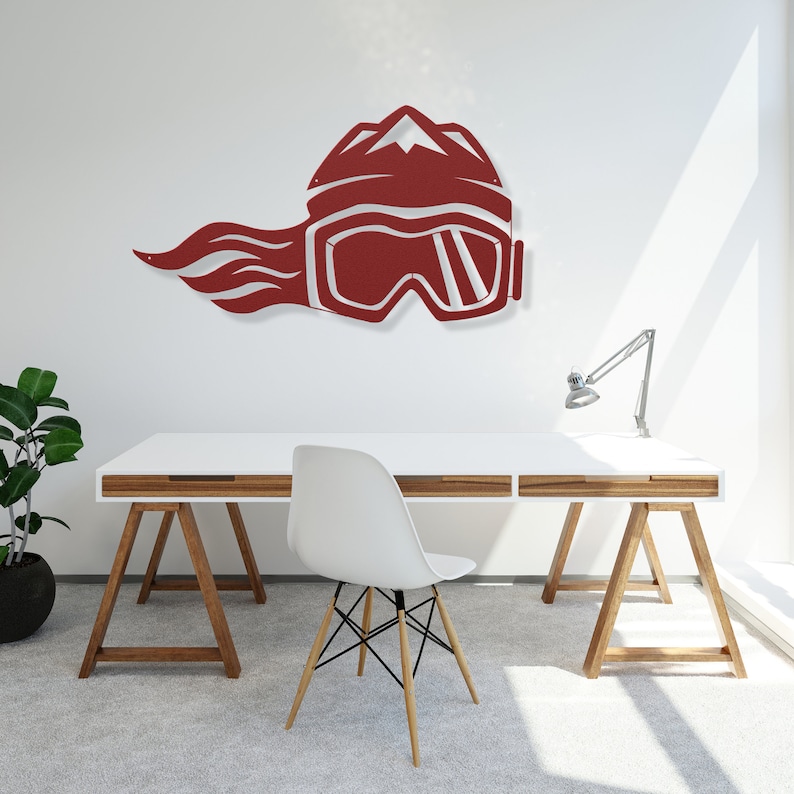 May include: A red metal wall art piece depicting a stylized ski helmet with goggles and a mountain peak design. The helmet has a flame-like design extending from the back. The art is mounted on a white wall.