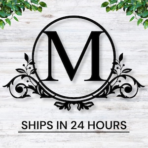 May include: Black metal monogram with a decorative floral design and the letter "M" in the center. The monogram is on a white wood background. "SHIPS IN 24 HOURS" is written below the monogram.