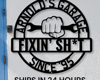 May include: Black metal sign with the text "Arnold's Garage Fixin' Sh*t Since '95" in a circle with a wrench and fist graphic.