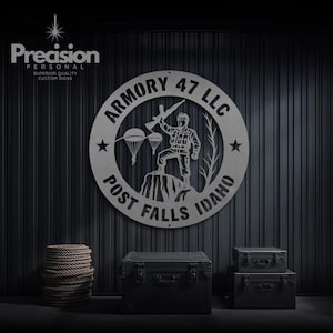 May include: A circular metal sign featuring the text ARMORY 47 LLC and POST FALLS IDAHO. The centre depicts a soldier standing on a rock formation holding a rifle, with two parachutes and a tree silhouette. Two stars flank the central design.