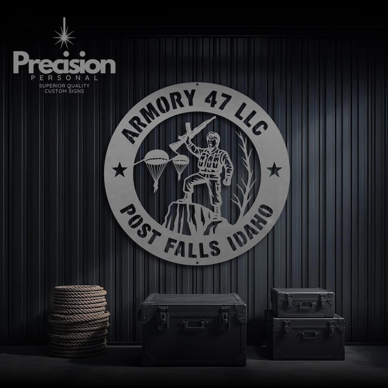 May include: A circular metal sign featuring the text ARMORY 47 LLC and POST FALLS IDAHO. The centre depicts a soldier standing on a rock formation holding a rifle, with two parachutes and a tree silhouette. Two stars flank the central design.
