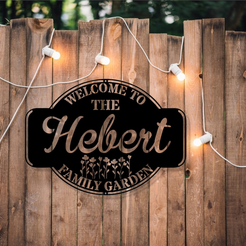 May include: Black metal sign with the text "Welcome to the Hebert Family Garden" in a decorative font. The sign is hanging on a wooden fence with string lights.