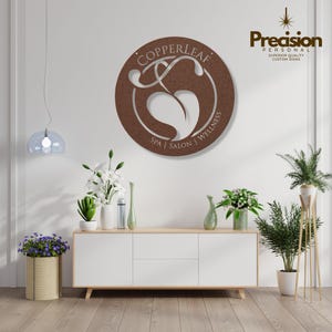 May include: A circular brown metal wall sign featuring the text Copperleaf Spa | Salon | Wellness with a central leaf cutout design. The sign is mounted on a white wall above a white cabinet with various potted plants and a hanging light fixture.