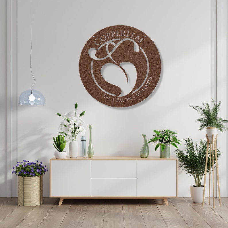 May include: A round, brown metal sign with the text "COPPERLEAF" and "SPA | SALON | WELLNESS" in white. The sign features a stylized leaf design. The sign is mounted on a white wall above a white cabinet with plants and flowers.