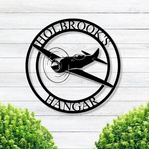 May include: Black metal sign with a circle design. The circle has the text "HOLBROOK'S HANGAR" around the outside and a silhouette of a plane inside the circle.