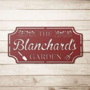 K&ouml;nnte beinhalten: Rotes Metall-Gartenschild mit dem Text "The Blanchard's Garden" mit einem Design aus Schaufel und Gabel.