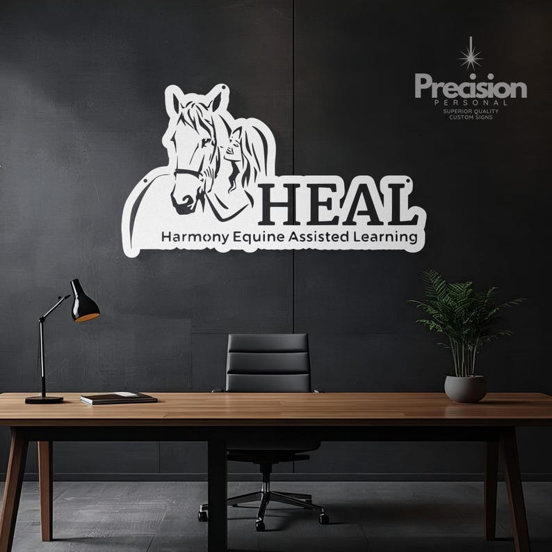May include: A white wall-mounted sign featuring a graphic of a horse and a person, with the text HEAL and Harmony Equine Assisted Learning. The sign is displayed on a dark wall above a wooden desk with a black lamp, a notebook, and a potted plant.