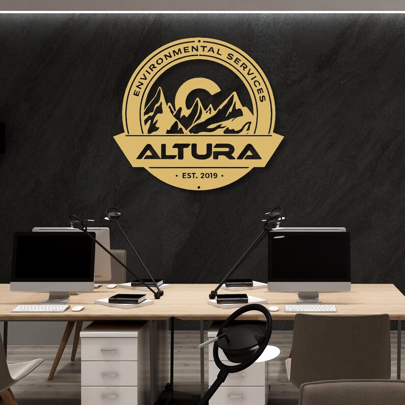 May include: A gold-colored sign with the text "ENVIRONMENTAL SERVICES" in a circular design, and "ALTURA" below it. The sign also includes the text "EST. 2019." The sign is mounted on a black wall above a desk with two computer monitors.