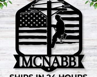 May include: Black metal silhouette of a lineman climbing a utility pole with an American flag background. The silhouette is in front of a wooden background. The text "MCNABB" is below the silhouette.