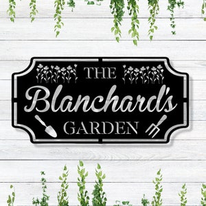 K&ouml;nnte beinhalten: Schwarzes Garten Schild aus Metall mit silberner Beschriftung "The Blanchard's Garden". Das Schild hat einen dekorativen Rand mit einer Schaufel und einer Heugabel auf beiden Seiten des Textes.