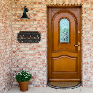 K&ouml;nnte beinhalten: Ein schwarzes Metallschild mit dem Text "The Blanchard's Garden" mit einem Spaten- und Rechen-Design an einer Backsteinwand neben einer Holzt&uuml;r mit einem Glasfenster.