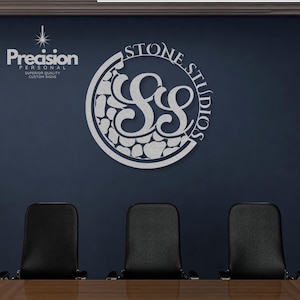 May include: A circular silver metal wall sign featuring the text STONE STUDIOS and a stylised SS monogram. The design includes a textured pattern resembling stacked stones within the circular frame. The sign is mounted on a dark blue wall.