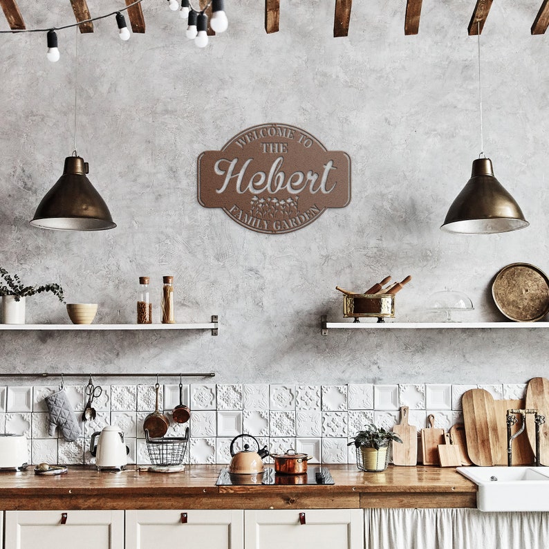 May include: A brown wooden sign with the text "Welcome to the Hebert Family Garden" in white lettering. The sign is hanging on a gray wall in a kitchen.