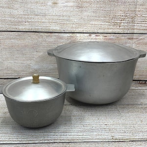 May include: Two aluminium cooking pots with lids. The larger pot has two side handles, and the smaller pot has a wooden knob on the lid. Both pots are on a wooden surface.