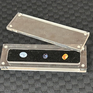 Puede incluir: Una vitrina de acrílico transparente con tapa abatible, que muestra tres piedras preciosas. Las piedras preciosas son azul claro, azul oscuro y naranja. La vitrina está sobre una superficie negra texturizada.