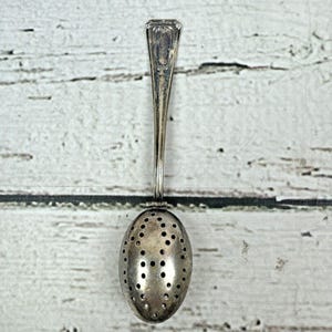May include: A vintage silver tea infuser spoon. The spoon has an oval-shaped bowl with numerous small holes and a long, slender handle. The handle has a decorative design at the top. The tea infuser is made of a shiny, metallic material.