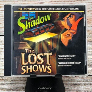 Puede incluir: Una caja de CD de "The Shadow: The Lost Shows" con una ilustración vintage. La portada presenta un cofre del tesoro, una figura sombría y el título del programa en verde. También se enumeran las fechas de emisión.