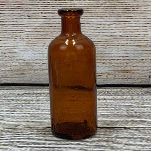 May include: A vintage brown glass bottle with a rounded body and a narrow neck. The bottle has a dark amber color and appears to be made of thick glass. The bottle is empty and sits on a wooden surface.