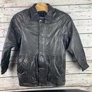May include: A black leather jacket with a collared neckline and button closures. The jacket features two front pockets and a buttoned cuff. The jacket is hanging on a wooden hanger against a white and gray wooden background.