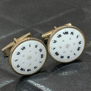 May include: Gold-coloured cufflinks with white faces, black Roman numerals, and star details. The round cufflinks have a watch face design. The image shows a close-up of the pair.