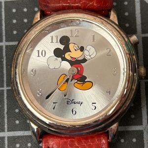 May include: A silver-toned wristwatch featuring Mickey Mouse on the face. The watch has a red leather band and a silver dial with black numerals. The Disney logo is printed below the center of the dial.