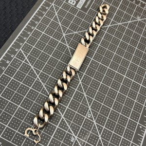 Puede incluir: Pulsera de cadena de eslabones plateada con una placa rectangular en el centro. La placa tiene las letras "FPM" grabadas. Un pequeño dije cuelga del cierre. La pulsera se muestra sobre una esterilla de corte gris.