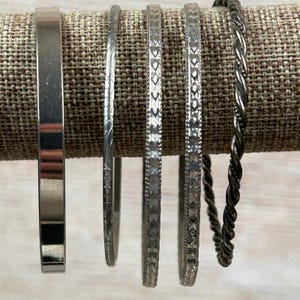 May include: A collection of silver-toned bangles, each with a unique design. The bangles include a smooth, flat band, a floral engraved band, and bands with geometric patterns. A twisted rope-style bangle is also included.