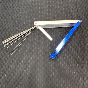 May include: A silver and blue carburetor jet cleaner tool. The tool has a silver handle with multiple thin silver wires extending from one end. A blue handle is attached to the other end.