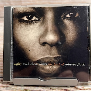 May include: A music CD in a clear plastic jewel case featuring a high-contrast, sepia-toned close-up portrait of a person's face. The text on the cover reads, softly with these songs the best of roberta flack, in white and orange lettering.