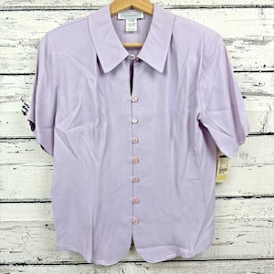 May include: A light purple short-sleeved blouse with a collared neckline and a series of small, round buttons down the front. The blouse has a slightly tailored fit and is hanging on a wooden hanger.