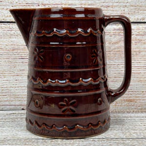 May include: A brown ceramic pitcher with a handle and spout. The pitcher has a glossy finish and is decorated with horizontal bands of wavy and floral patterns. The pitcher is approximately 9 inches tall.