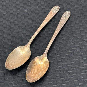 Puede incluir: Dos cucharas de plata antiguas con diseños florales ornamentados en los mangos. Las cucharas tienen cuencos ovalados y muestran signos de uso. El fondo es una superficie oscura y texturizada.