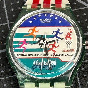Puede incluir: Un reloj Swatch con una caja verde transparente y una correa a rayas rojas y blancas. La esfera del reloj presenta el texto "swatch SWISS" y "Atlanta 1996". Tiene un fondo azul con estrellas blancas y corredores de varios colores.