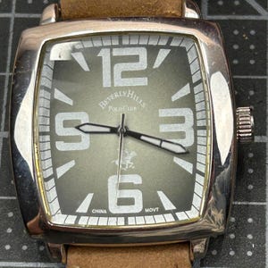 May include: A square-faced wristwatch with a silver-tone case and a brown leather strap. The watch face is olive green with large white numerals at 12, 3, 6, and 9 o'clock. The brand name "Beverly Hills Polo Club" is displayed.