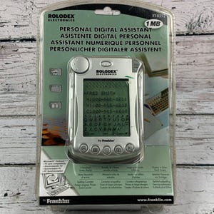 May include: A silver Rolodex Personal Digital Assistant, model RT-8212, in its original packaging. The screen displays contact information, including a name and phone numbers. The packaging includes the Franklin brand name and product features.