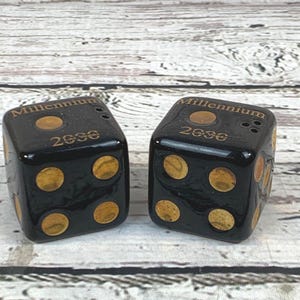 Könnte beinhalten: Zwei schwarze Würfel mit goldfarbenen Punkten und dem Text "Millennium 2000". Die Würfel sind quadratisch und glänzend. Sie liegen auf einer hellen Holzoberfläche.
