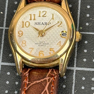 May include: A gold-coloured Sharp quartz watch with a light-coloured face, featuring black numerals and hands. The watch has a brown leather strap. The face reads "SHARP" and "100FT WATER RESIST QUARTZ".