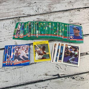 Puede incluir: Una colección de cromos de béisbol con varios jugadores. Las tarjetas tienen bordes verdes, azules y amarillos, con imágenes de jugadores y logotipos de equipos. Algunas tarjetas están apiladas, mientras que otras están abanicadas.