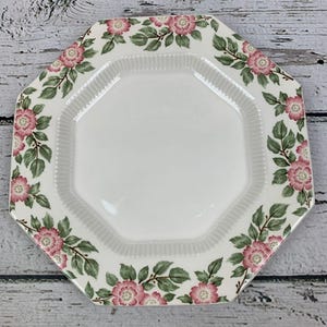 May include: An octagonal white ceramic plate with a floral design. Pink roses and green leaves decorate the border. The plate has a ridged inner edge and a smooth, white center. A decorative piece for serving.