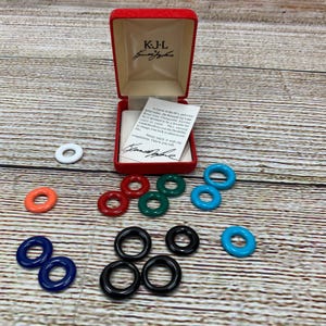May include: A selection of colourful, round earrings in shades of white, orange, red, green, blue, and black. The earrings are arranged on a wooden surface, with a red jewellery box bearing the initials "KJL" and a signature.