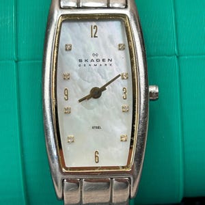 Puede incluir: Un reloj rectangular Skagen Denmark con una correa de acero inoxidable plateado. La esfera es blanca con detalles dorados, con los números 12, 3, 6 y 9, y pequeños marcadores cuadrados. La palabra "STEEL" está impresa en la parte inferior.