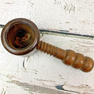 Könnte beinhalten: Ein Vintage-Nussknacker aus Holz mit einer runden Schüssel und einem geriffelten Griff. Der Nussknacker ist aus dunkelbraunem Holz gefertigt und zum Knacken von Nüssen bestimmt. Der Griff hat eine Reihe von abgerundeten Abschnitten für den Halt.