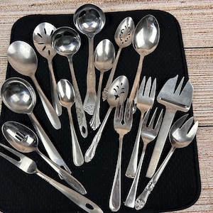May include: A collection of vintage silverware, including spoons, ladles, forks, and serving utensils. The silver-toned pieces are arranged on a black mat against a light wood background. Various designs and sizes are visible.